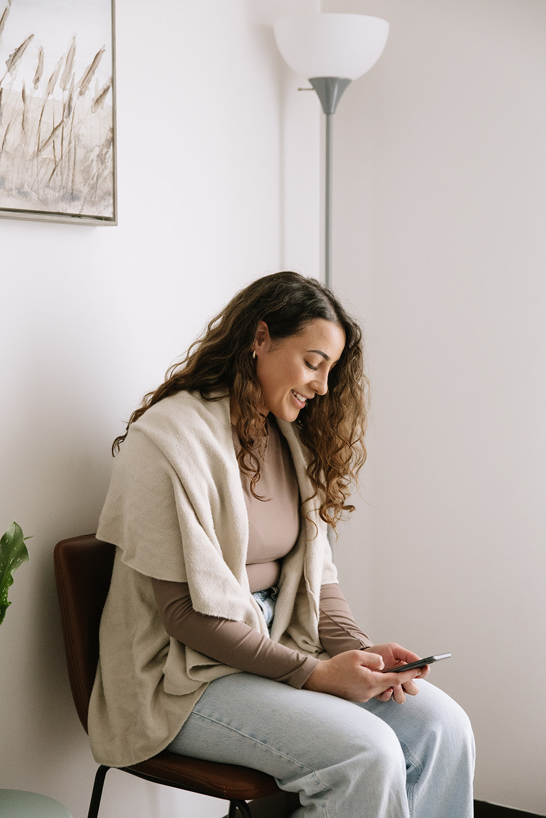 A woman sitting comfortably in a bright room, smiling as she looks at her phone, symbolizing self-connection, independence, and the empowered shift away from people-pleasing patterns at The Mindful Place in Ontario.