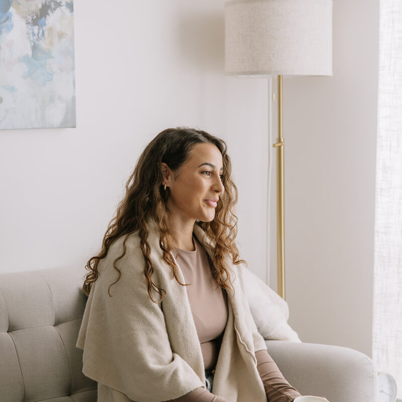 A woman sitting on a light-coloured couch with a warm drink, smiling gently in a calm, bright room, symbolizing comfort, safety, and the supportive process of beginning therapy at The Mindful Place in Ontario.