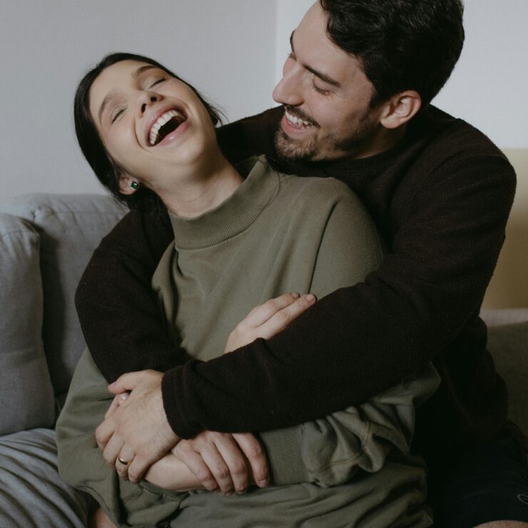 A couple sitting together on a couch, laughing and holding one another warmly, reflecting closeness, emotional safety, and the exploration of attachment patterns in relationships, at The Mindful Place in Ontario.